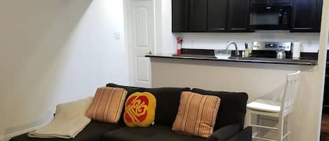 Ein Wohnzimmer mit Ecksofa, eine Küche mit dunklen Schränken und einer weißen Tür.