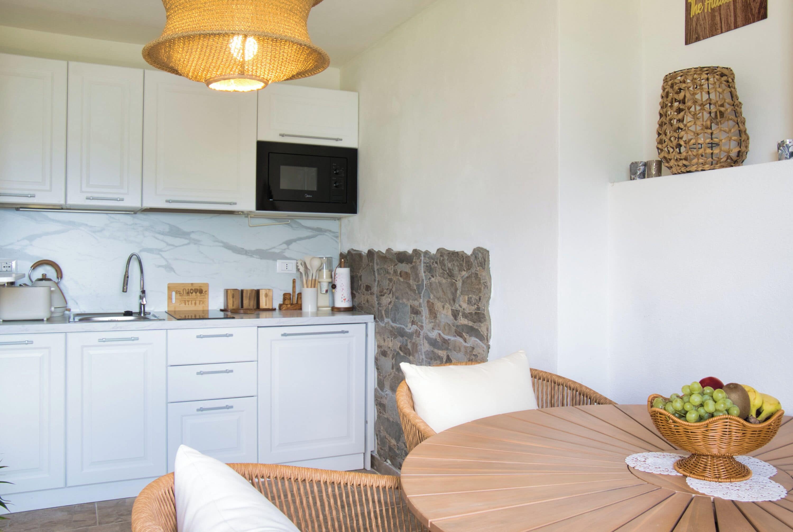 Cocina con gabinetes blancos, microondas y pared de piedra. Hay una mesa de mimbre con un frutero y un mantel individual blanco.