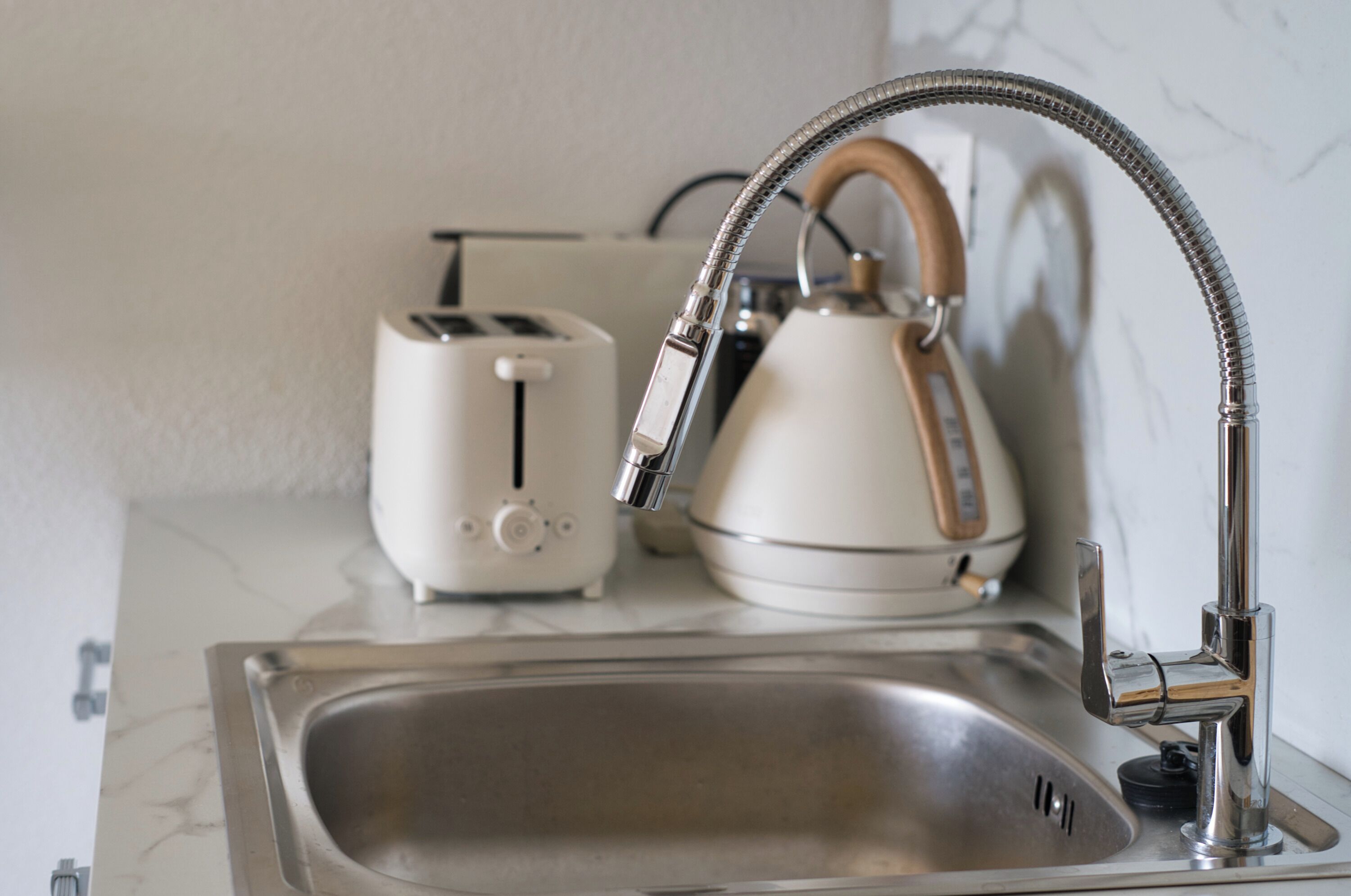 A kitchen with a stainless steel sink, a white toaster, and a white kettle with a wooden handle.