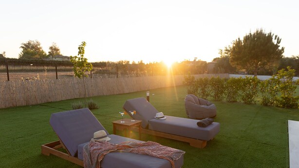 A sunlit outdoor seating area with two lounge chairs and a bean bag chair, a small table with a drink, and a blanket on the grass.
