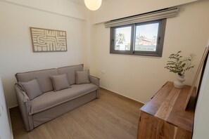 A room with a beige sofa, a wooden side table with a vase, a window with blinds, and a wall-mounted artwork.