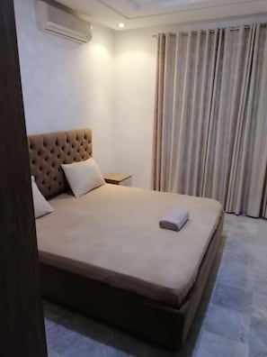 Ein Hotelzimmer mit einem großen Bett, einem Nachttisch, einer Klimaanlage und einem Vorhang.