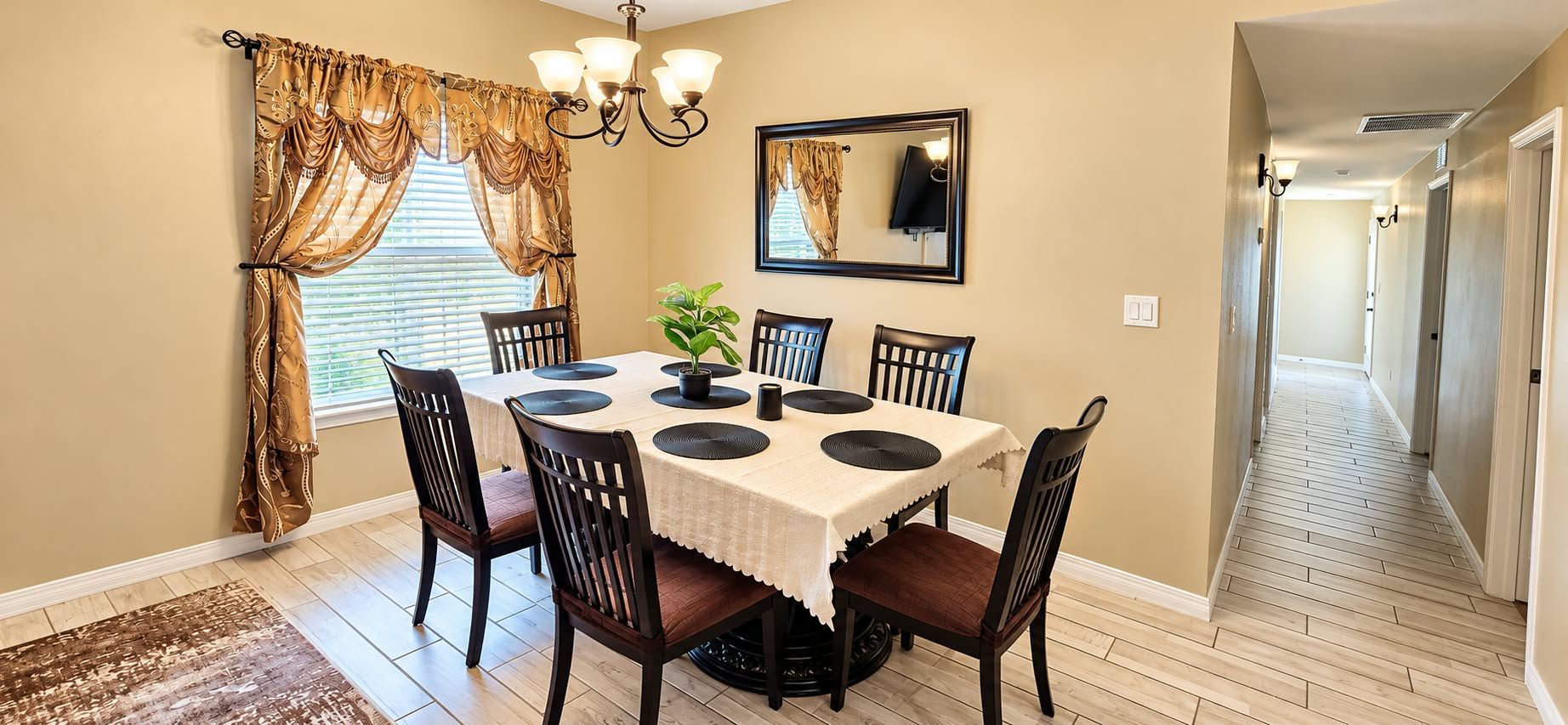Un coin repas avec une table dressée pour six personnes, entourée de chaises, d’un lustre et d’un miroir au mur.
