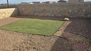 A backyard with a square area of artificial grass, surrounded by a stone wall and gravel ground.