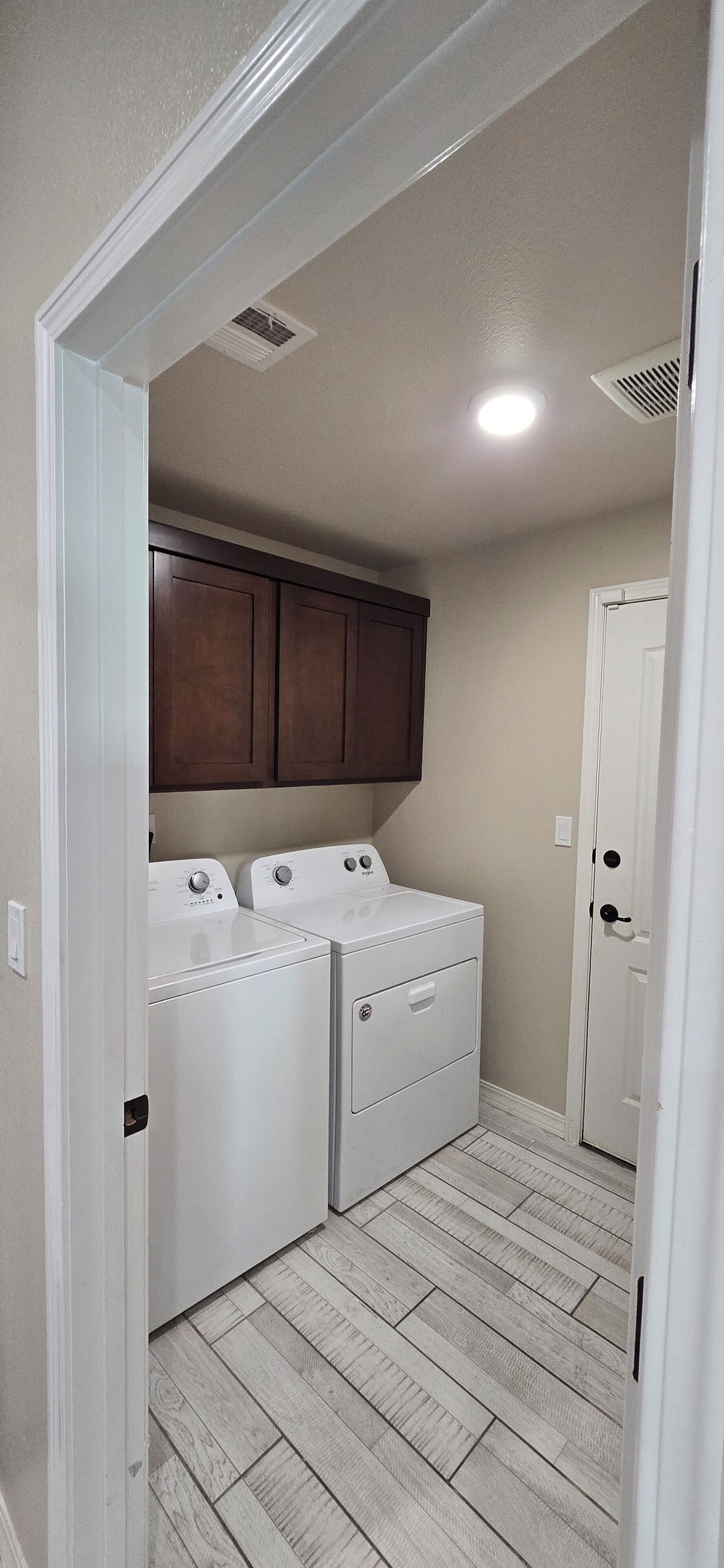 A laundry room with two white front-loading washers and dryers, wooden cabinets above, and a door to the right.