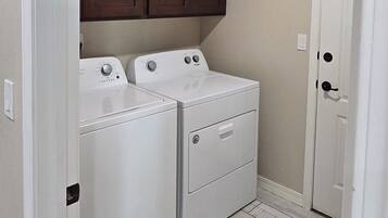 A laundry room with two white front-loading washers and dryers, wooden cabinets above, and a door to the right.