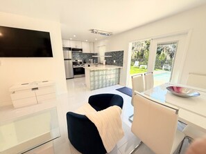 A modern kitchen with a large island, a dining area with a glass table, and a view of a pool through the window. - Short Walk to Beach • 3BR Poolside Unit • Next to Nature Preserve (Fort Myers Beach)