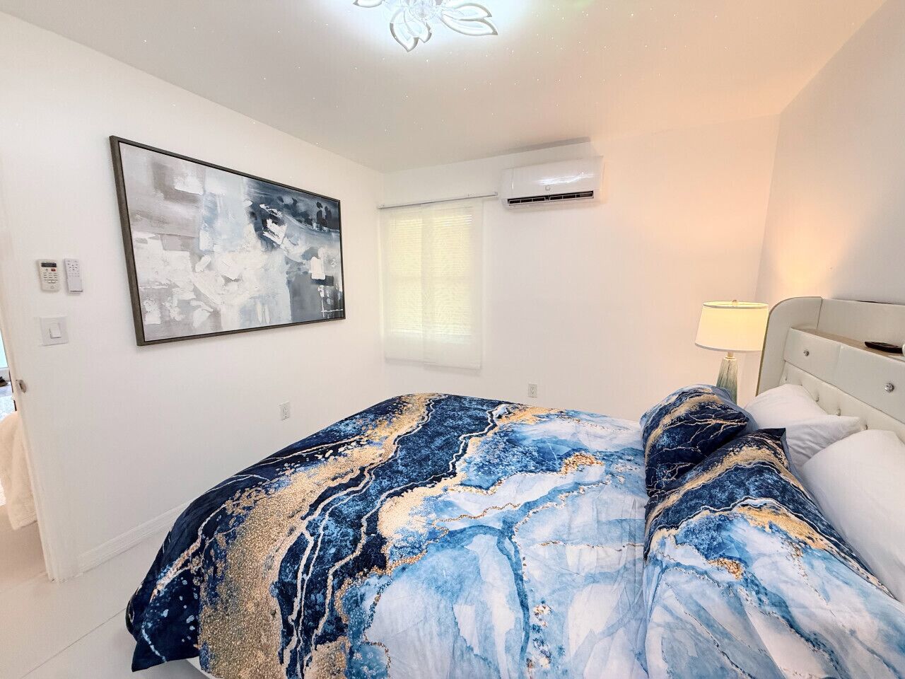 A bedroom with a bed, a wall-mounted air conditioner, a window with blinds, and a painting on the wall.
