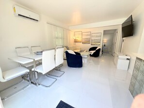 A modern living room with a dining area, a flat-screen TV mounted on the wall, and a large window with curtains. - Big Groups,Nature View Estates 3 units private pool, WALK TO BEACH (Fort Myers Beach)