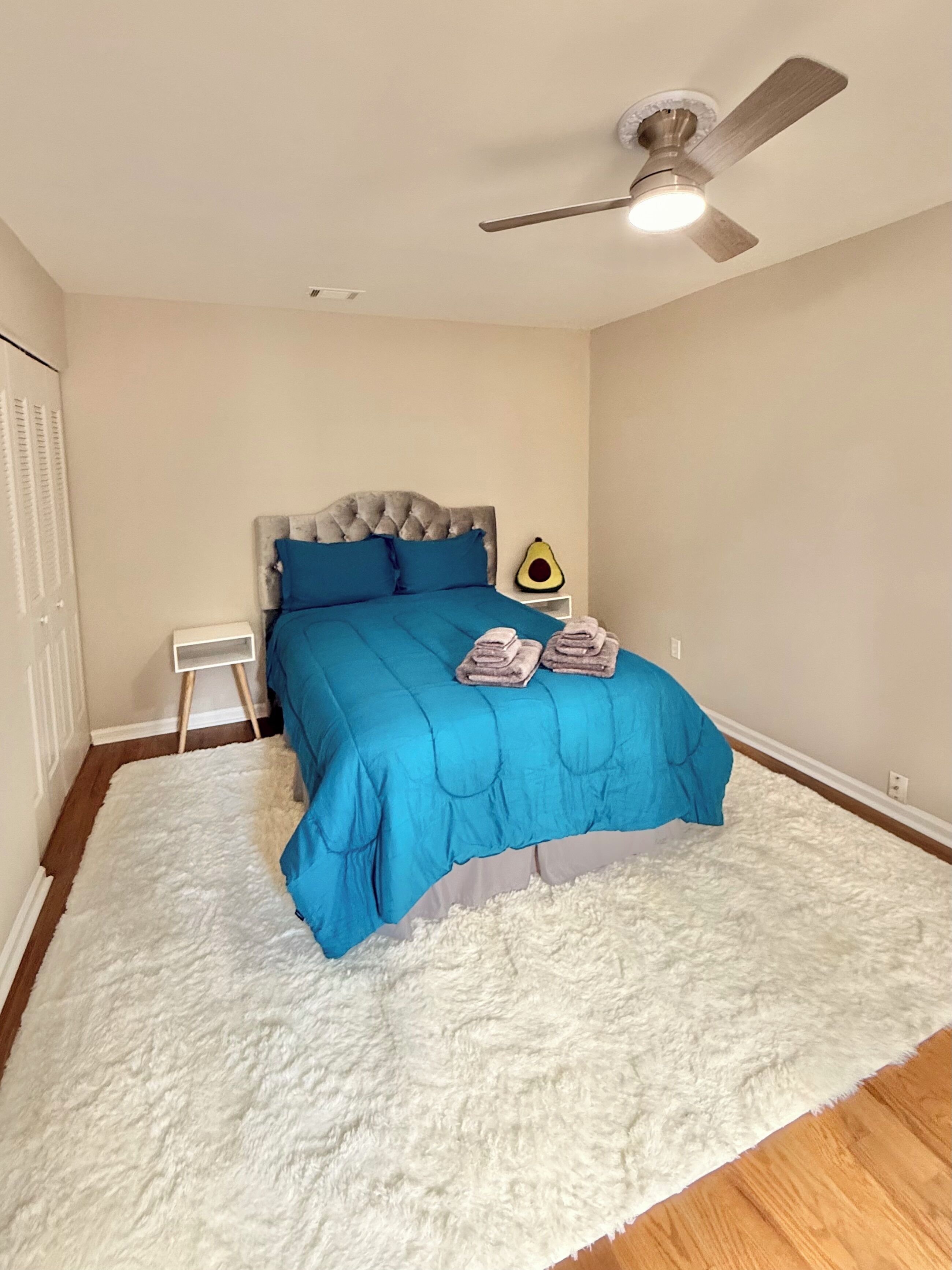 A bedroom with a bed, a ceiling fan, a nightstand, and a white fluffy rug.