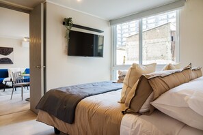 Ein modernes Schlafzimmer mit einem Bett, einem an der Wand befestigten Fernseher und einem Fenster mit Blick auf Gebäude.
