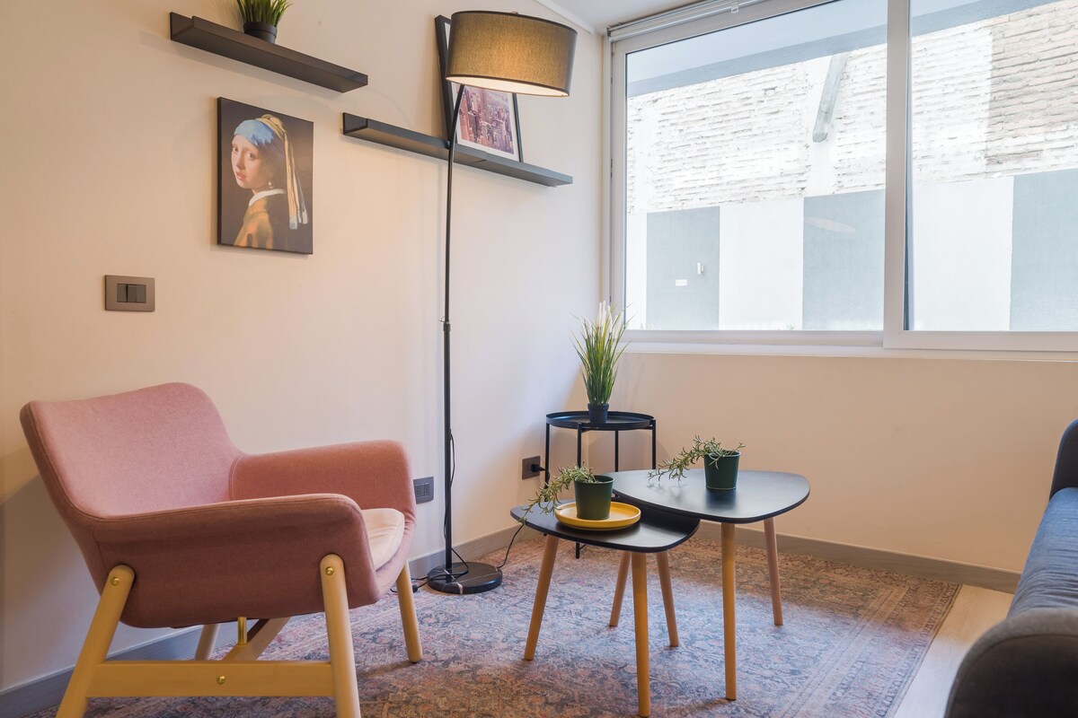 A modern living room with a pink armchair, a floor lamp, a small table with potted plants, and a large window.