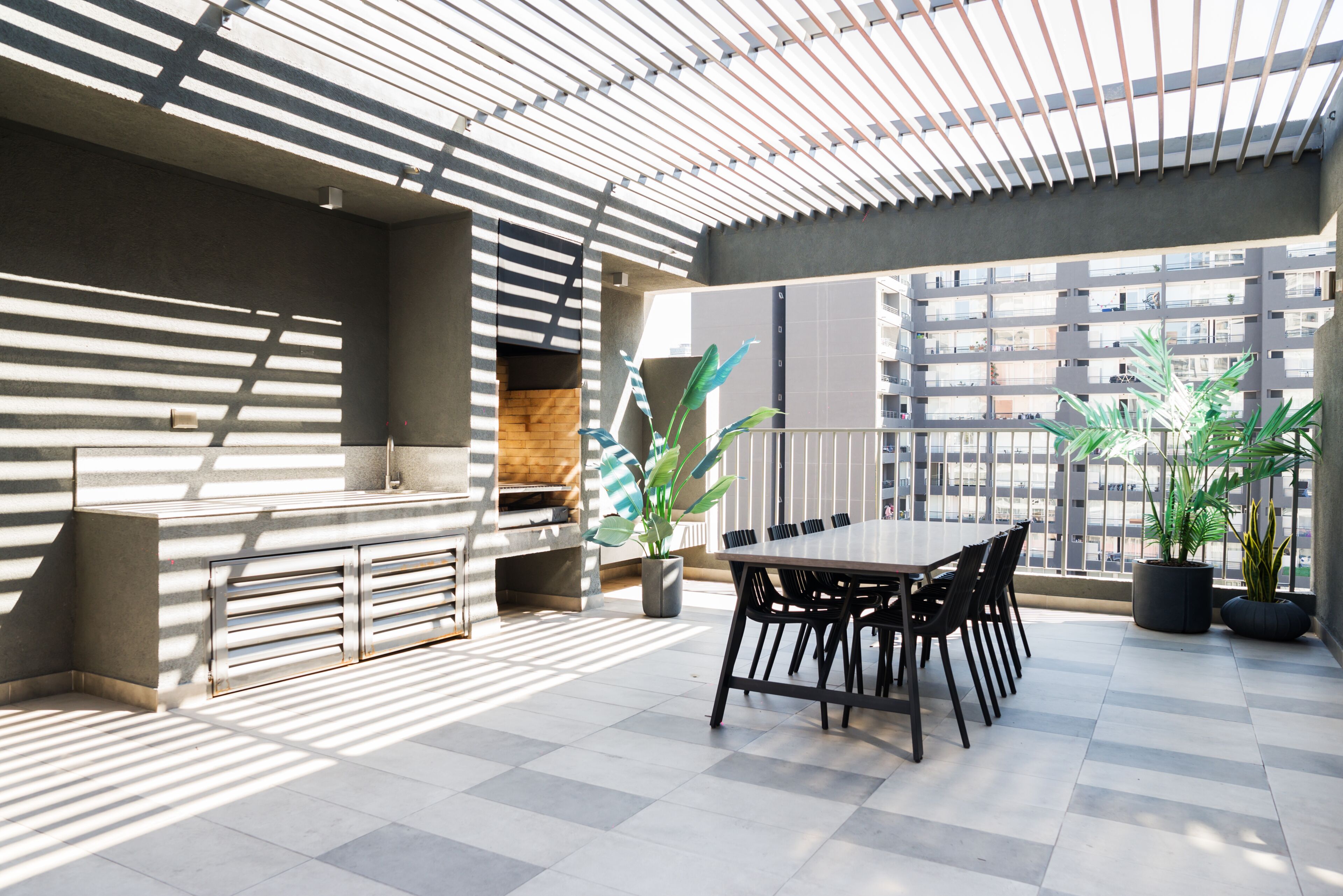Un espacio exterior moderno para comer con una mesa y sillas, rodeado de plantas en macetas y vistas a un edificio de gran altura.