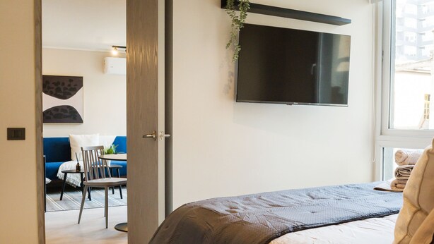 A modern bedroom with a bed, a TV mounted on the wall, and a door leading to a dining area. - Eleuterio Ramirez Apartments by Andes (Santiago)