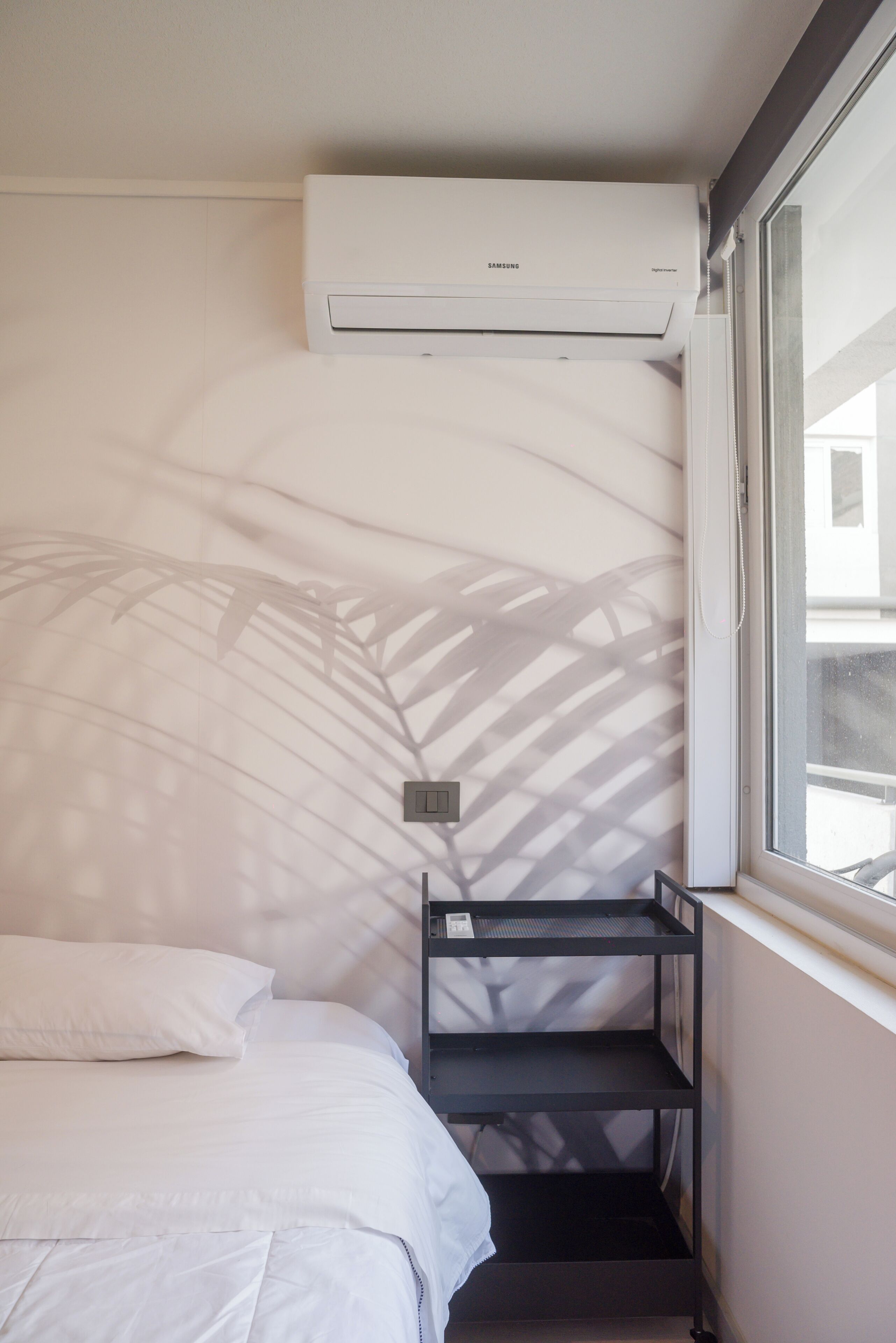 A hotel room with a bed, a small shelf, an air conditioning unit, and a window with a view.