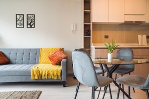 Ein modernes Wohnzimmer mit einem grauen Sofa, einem gelben Überwurf und orangefarbenen Kissen. Es gibt einen Glastisch mit zwei Stühlen und einer Topfpflanze.