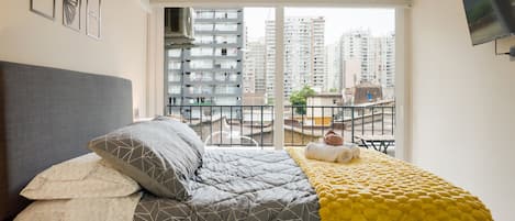 Een slaapkamer met een bed, een raam en uitzicht op hoge gebouwen.