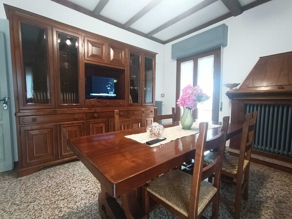 A dining room with a wooden table, chairs, a television on a cabinet, and a vase with flowers.