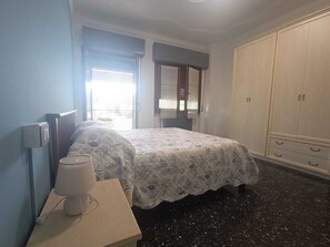 Ein Schlafzimmer mit Bett, Nachttisch mit Lampe, Kleiderschrank und einem Fenster mit Vorhängen.