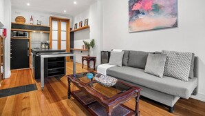 A modern living room with a grey sofa, a coffee table, and a kitchen area with wooden cabinets and a microwave. - Rosa and Noir Riverside Escape near Botanic Gardens (South Yarra)
