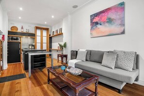 A modern living room with a grey sofa, a coffee table, and a kitchen area with wooden cabinets and a microwave. - Rosa and Noir Riverside Escape near Botanic Gardens (South Yarra)