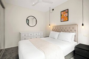 Une chambre à coucher comprenant un lit, une commode blanche, un miroir rond, un ventilateur de plafond et un luminaire fixé au mur.