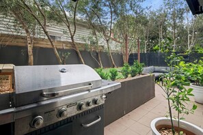 Ein Außenbereich mit einer Terrasse, einem Grill aus Edelstahl, einem Betonpflanzgefäß mit einem kleinen Baum und einem grauen Outdoor-Sofa.
