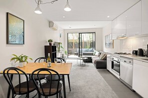 A modern kitchen with white cabinets, a dining area with a wooden table and chairs, and a living area with a sofa and a coffee table. - Richmond Oasis Private Courtyard near MCG and Cafs (Richmond)