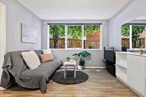 Un espace de vie compact comprenant un canapé, une petite table, un téléviseur et une fenêtre donnant sur une clôture en bois et de la verdure.