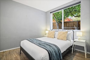 A bedroom with a bed, bedside tables, and a window with a view of a wooden fence and trees. - Richmond Pad MCG and Bridge Rd Stay Free Parking (Richmond)
