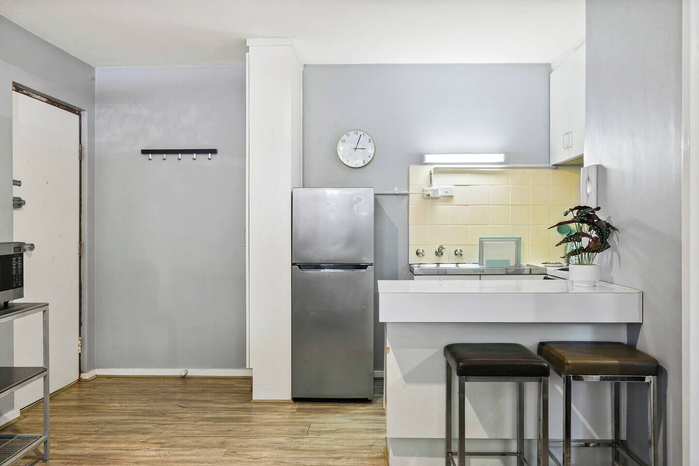 A compact kitchen with a stainless steel refrigerator, a microwave, a sink with a mirror, and a clock on the wall. There are two stools and a potted plant on the counter.