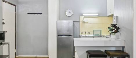 A compact kitchen with a stainless steel refrigerator, a microwave, a sink with a mirror, and a clock on the wall. There are two stools and a potted plant on the counter.