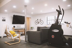 Ein Wohnzimmer mit Flachbildfernseher, einer Couch, einem Sessel mit Decke sowie einem Fahrrad und einem Heimtrainer.