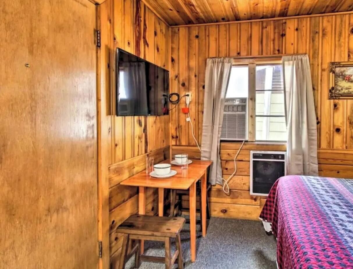 A wooden cabin room with a bed, a table with a chair, a television mounted on the wall, and a window with curtains.