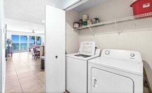 A laundry room with two white washing machines, a shelf with cleaning supplies, and a view of a balcony with a table and chairs. - Oceanfront Condo with Shared Pool & Fitness Room (Destin)