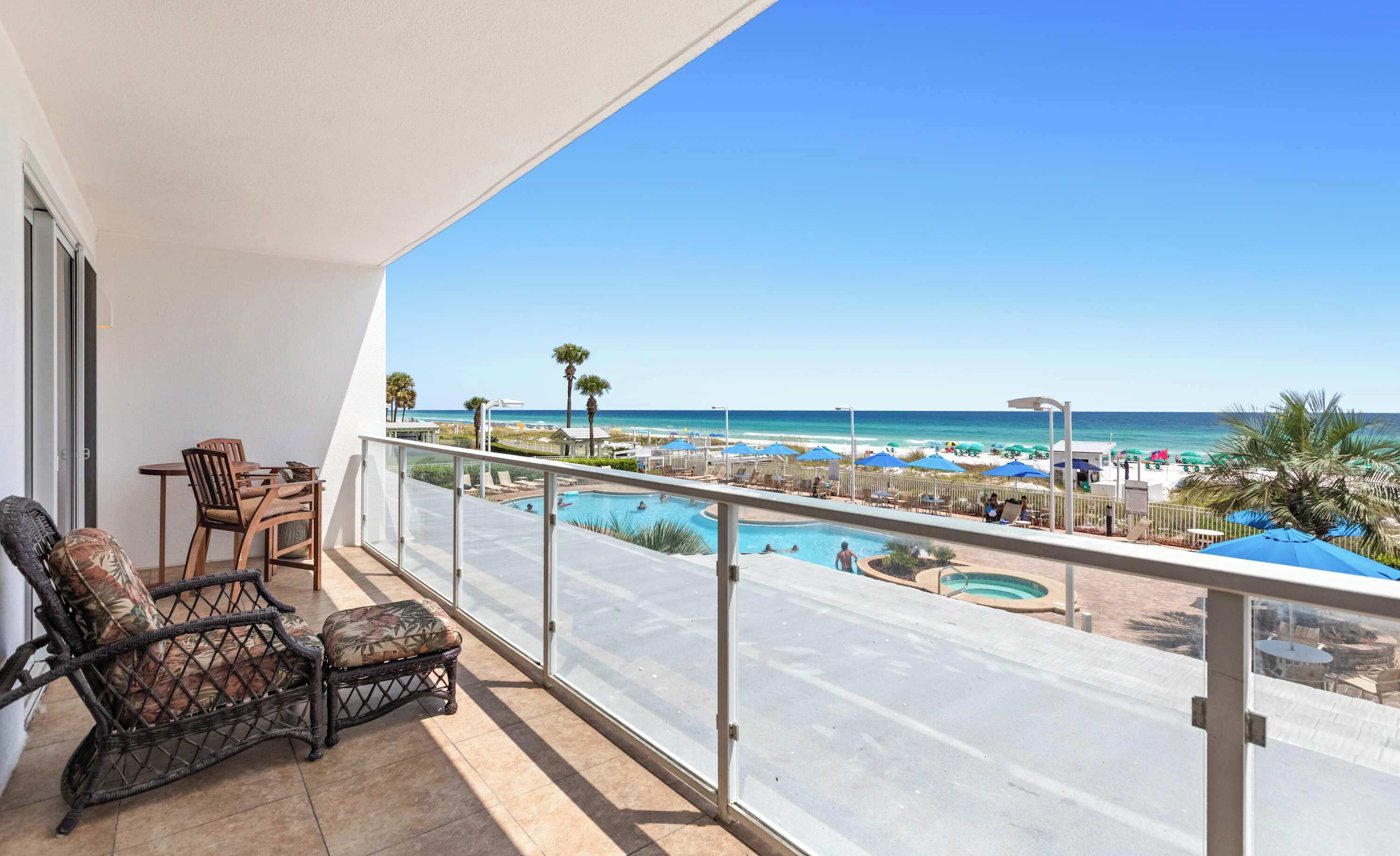 A balcony with a view of a beach, a pool, and lounge chairs.