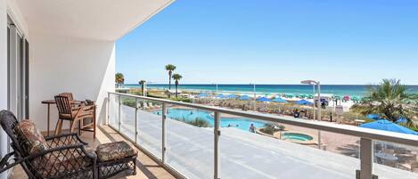 A balcony with a view of a beach, a pool, and lounge chairs.