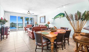 A dining area with a wooden table and chairs, a ceiling fan, and a view of the ocean through large windows. - Oceanfront Condo with Shared Pool & Fitness Room (Destin)