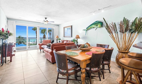 A dining area with a wooden table and chairs, a ceiling fan, and a view of the ocean through large windows. - Oceanfront Condo with Shared Pool & Fitness Room (Destin)