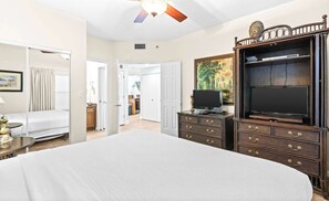 A hotel room with a large bed, a wardrobe with a mirror, a television, and a ceiling fan. - Oceanfront Condo with Shared Pool & Fitness Room (Destin)