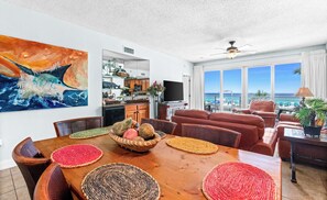 A spacious living area with a wooden dining table, red woven placemats, and a large painting of a fish. - Oceanfront Condo with Shared Pool & Fitness Room (Destin)