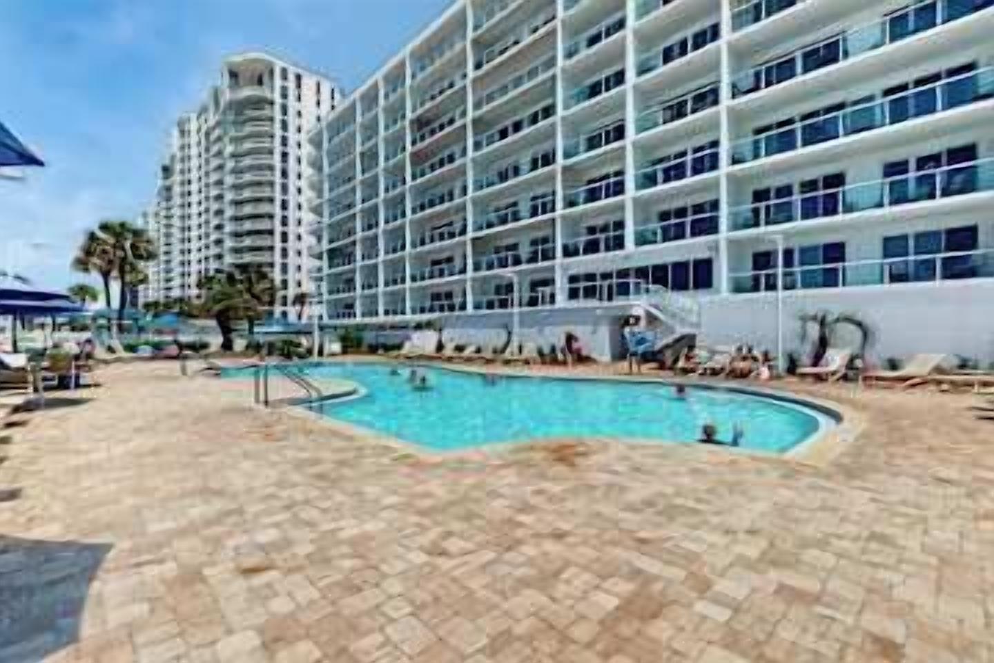 A hotel with a swimming pool, multiple balconies, and a large building in the background.