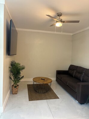 A living room with a dark brown sofa, a round wooden coffee table, a ceiling fan, a mounted flat-screen TV, and a potted plant. - Cozy 1-bedroom house in wonderful Nassau with AC and outdoor fitness area (Nassau)