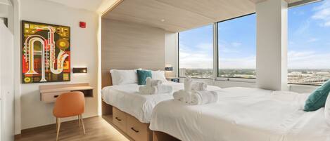 A modern hotel room with a large bed, a desk, a chair, and a city view through large windows.