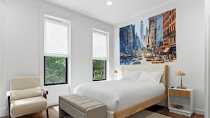 A bedroom with a large bed, a chair, a bench, a nightstand, and a painting of a cityscape.