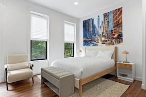 A bedroom with a large bed, a chair, a bench, a nightstand, and a painting of a cityscape.