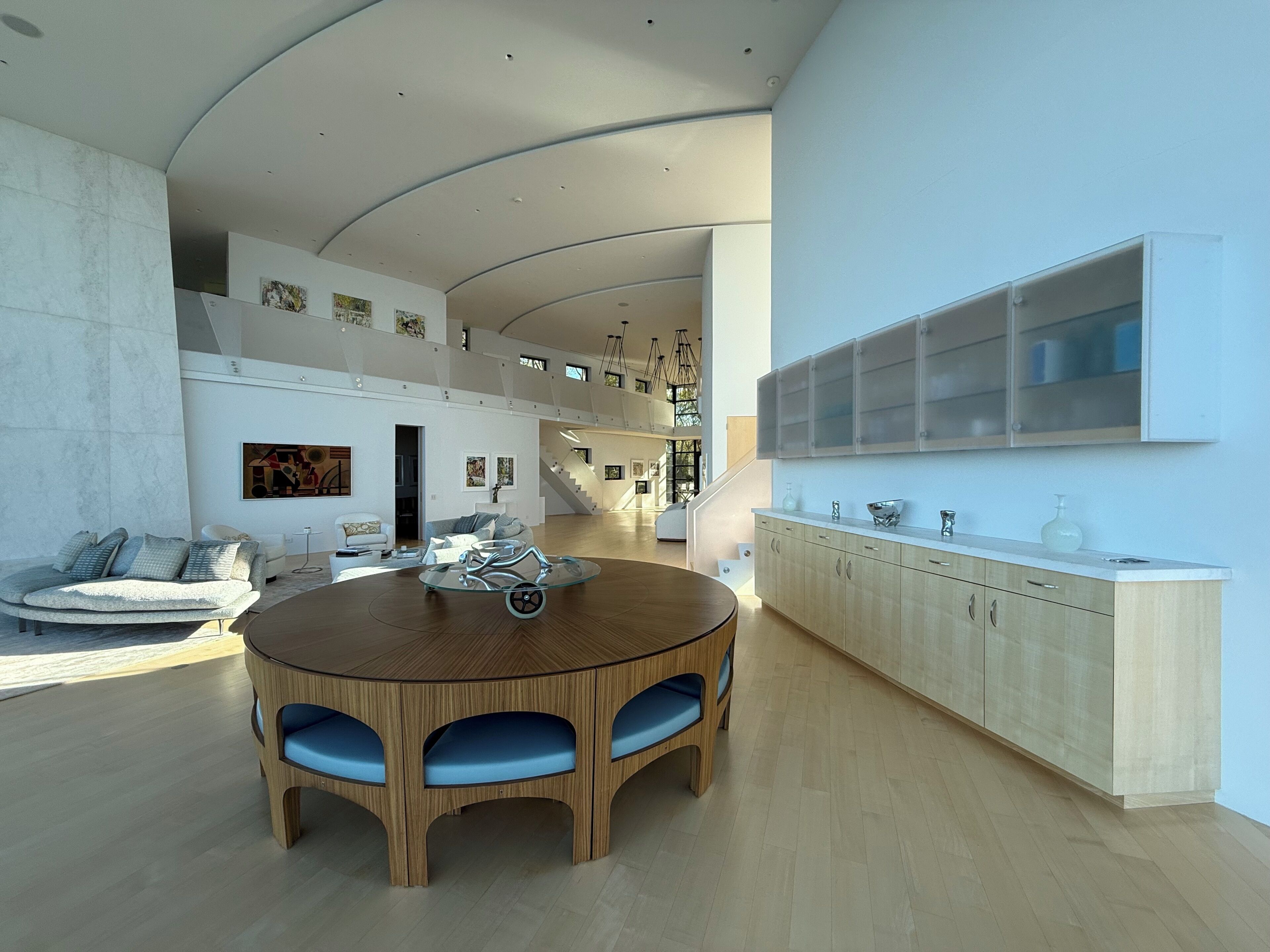 A modern interior with a round wooden dining table, blue cushioned chairs, a white sofa, and a staircase leading to an upper level.
