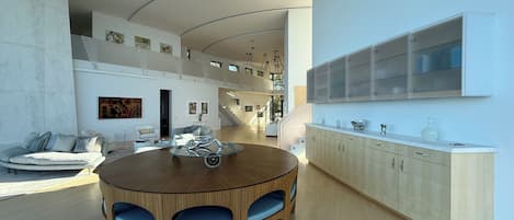 A modern interior with a round wooden dining table, blue cushioned chairs, a white sofa, and a staircase leading to an upper level.