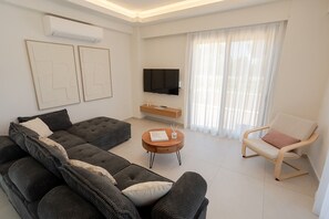Un salon moderne avec un canapé d’angle, une table basse ronde, un fauteuil blanc, un téléviseur à écran plat et une grande fenêtre ornée de rideaux légers.