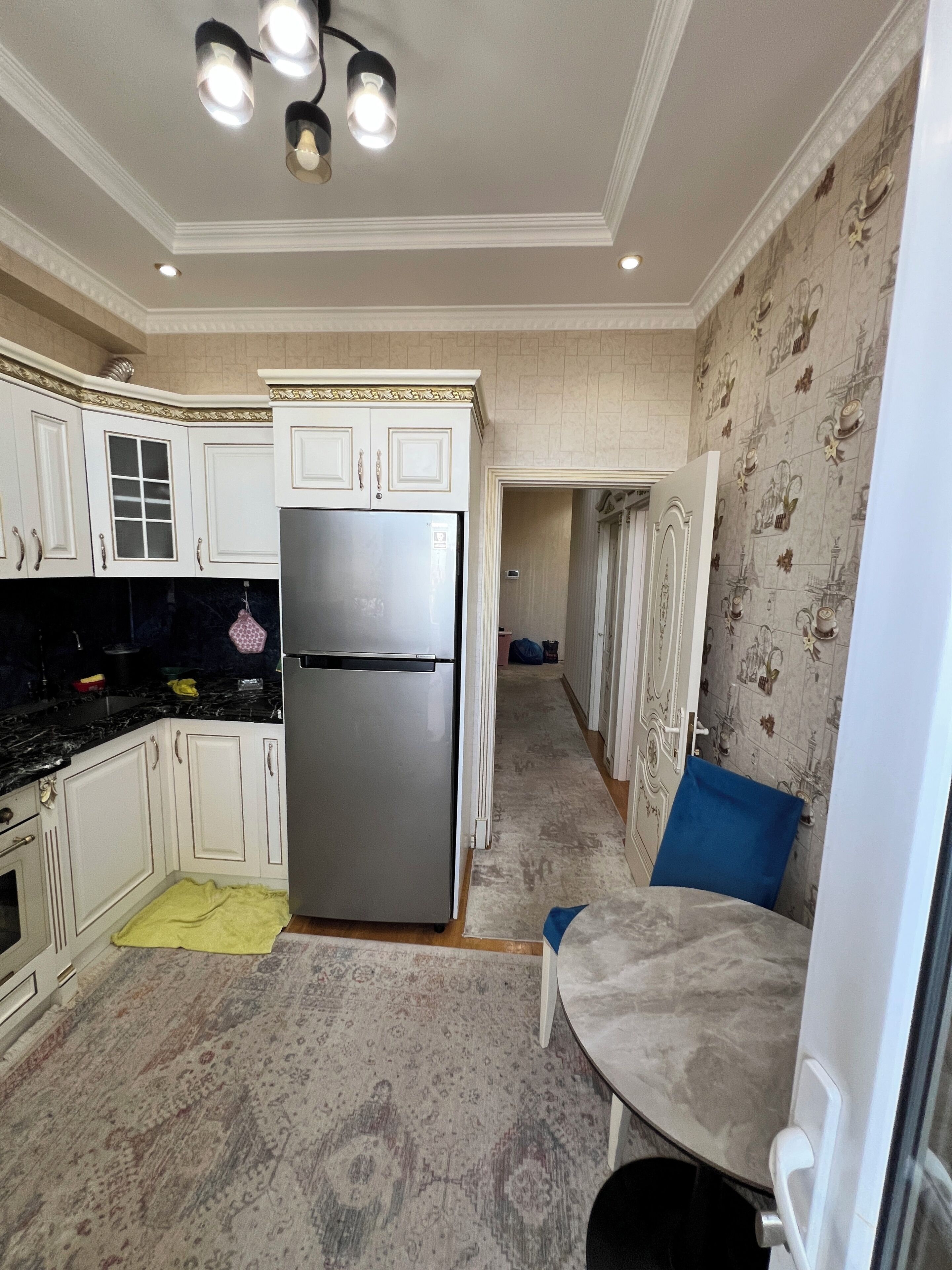 A kitchen with white cabinets, a stainless steel refrigerator, a patterned rug, and a blue chair.
